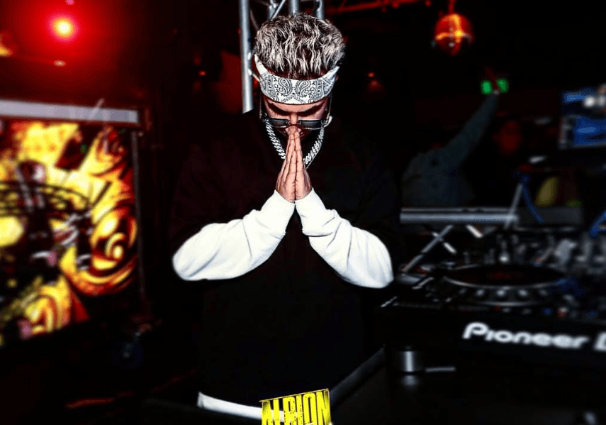 Artist DJ Saint in a club performance, hands in prayer pose, under neon lights.