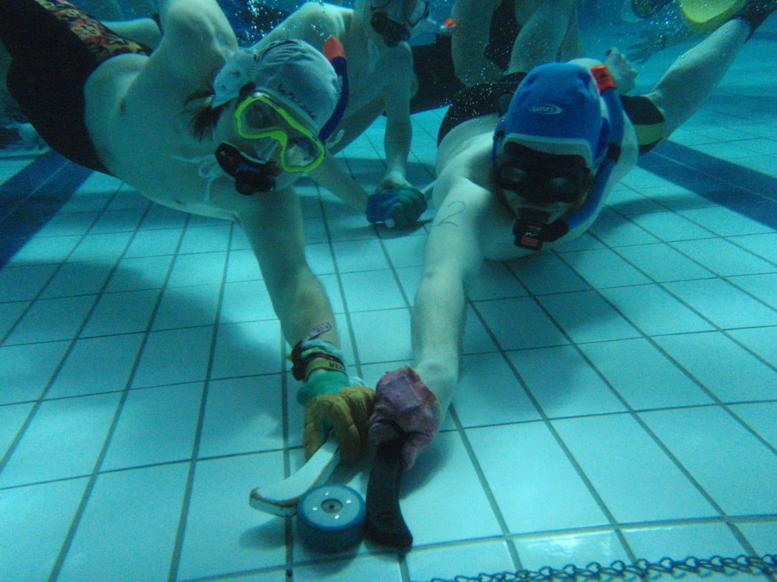 Two underwater hockey players reach for puck on tiled pool floor