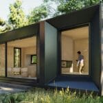 A modern prefab tiny house with glass doors and wood siding amidst green forest.